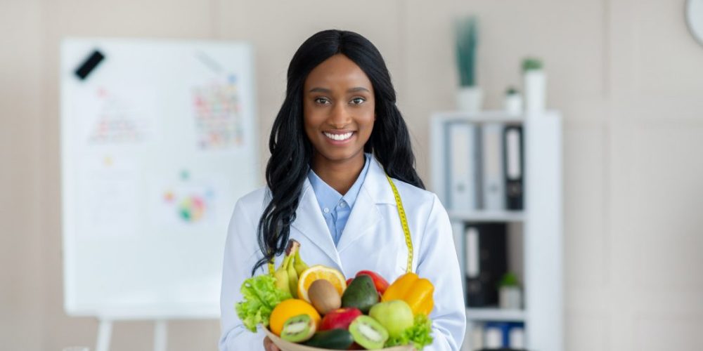 portrait-of-happy-black-dietitian-in-lab-coat-holding-bowl-of-fresh-fruits-and-vegetables-smiling